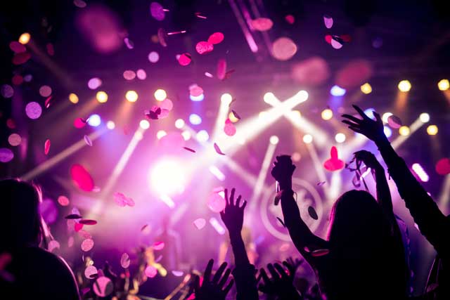 Eine Nahaufnahme einer feiernden Menge bei einem Konzert oder Festival, die die Hände in die Luft streckt, während Konfetti unter buntem Bühnenlicht herabregnet.